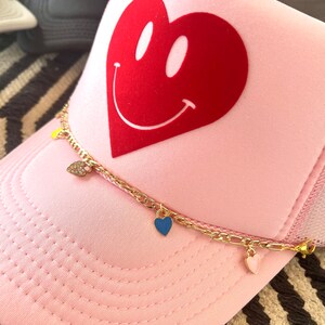 May include: Pink trucker hat with a red heart with a smiley face on it. The hat has a gold chain with three heart charms, one blue, one pink, and one clear.