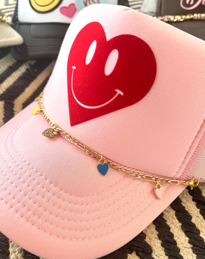 May include: Pink trucker hat with a red heart with a smiley face on it. The hat has a gold chain with three heart charms, one blue, one pink, and one clear.