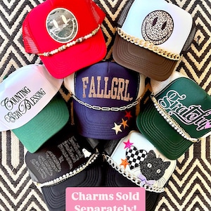 May include: A collection of seven trucker hats with various designs and embellishments. The hats feature a variety of colors, including red, white, green, black, blue, and brown. The hats are decorated with charms, beads, and chains. The text "Charms Sold Separately!" is displayed in pink at the bottom of the image.