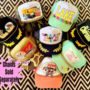 May include: A selection of trucker caps with diverse designs and text, such as "Lake Babe," "Lake," and "Girls Just Want to Have Fun." The caps have mesh backs, embroidered patches, and decorative chains.