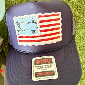 May include: Navy blue trucker hat with a patch featuring a stylized American flag design. The flag has red and white stripes and a blue floral pattern in the corner. The hat includes a snap closure and the text "OTTO TRUCKER".