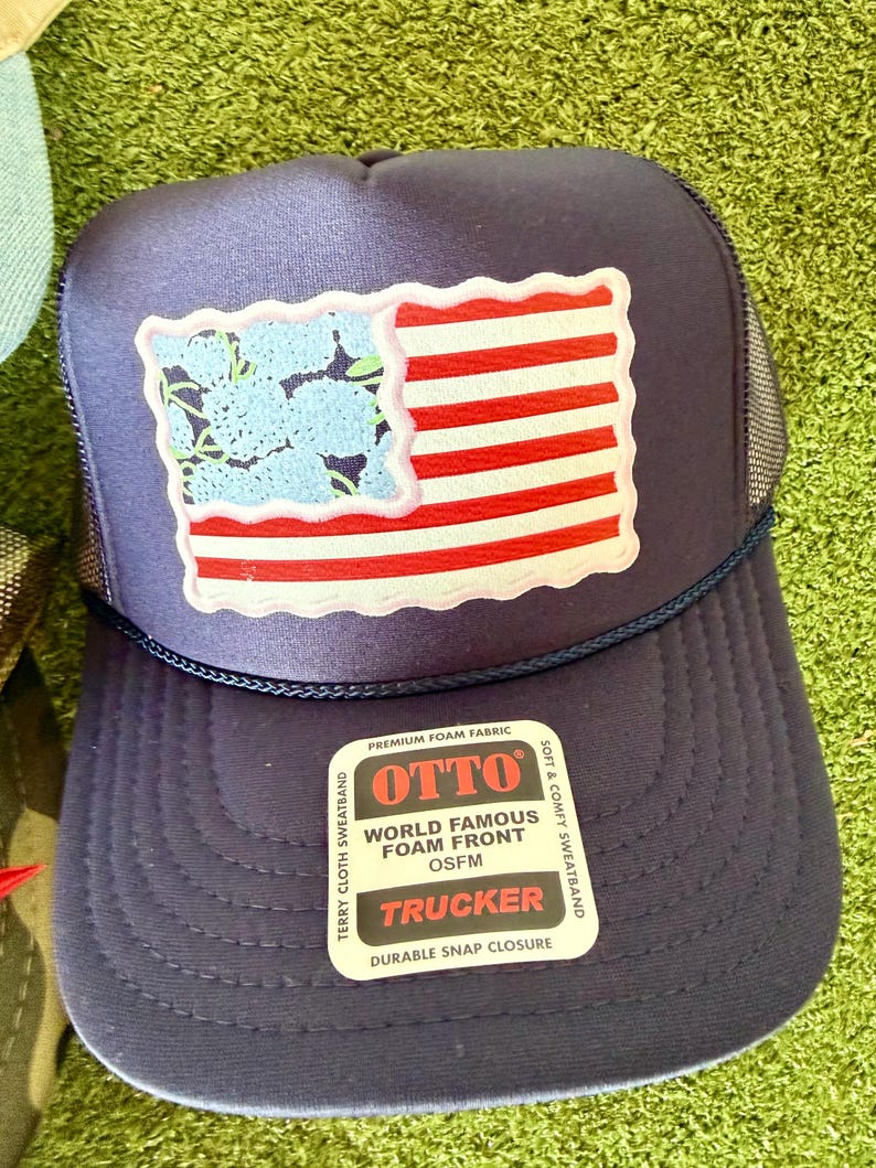 May include: Navy blue trucker hat with a patch featuring a stylized American flag design. The flag has red and white stripes and a blue floral pattern in the corner. The hat includes a snap closure and the text "OTTO TRUCKER".