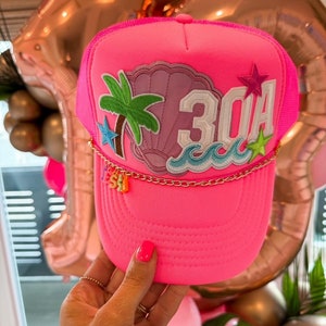 May include: Pink trucker hat with a seashell, palm tree, and waves design. The hat also has a gold chain with charms that spell out "SEA" and the number "30A" on the front.