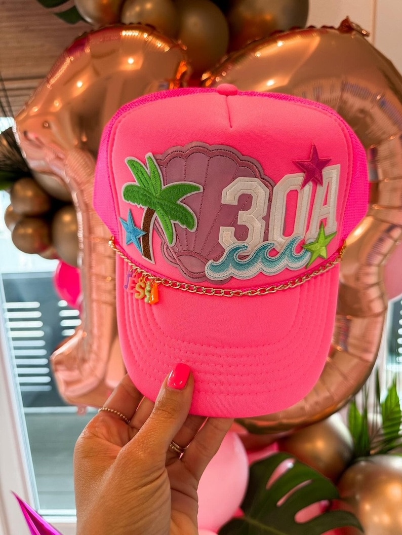 Op de afbeelding: Roze truckercap met een schelp, palmboom en golven ontwerp. De pet heeft ook een gouden ketting met bedels die "SEA" spellen en het nummer "30A" aan de voorkant.