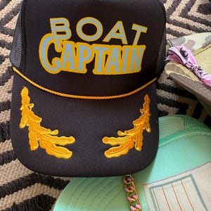 May include: Black trucker hat with the words "BOAT CAPTAIN" in blue and yellow lettering. The hat features gold embroidered leaf designs and a gold rope detail. Other hats are visible in the background.