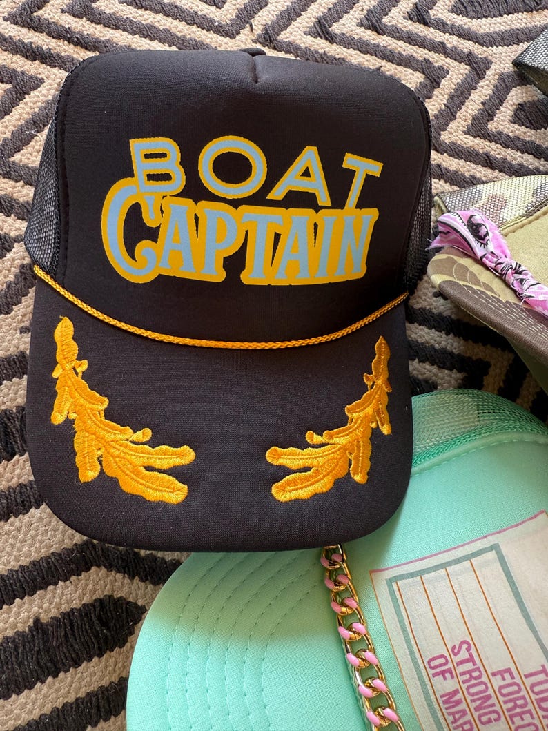 May include: Black trucker hat with the words "BOAT CAPTAIN" in blue and yellow lettering. The hat features gold embroidered leaf designs and a gold rope detail. Other hats are visible in the background.