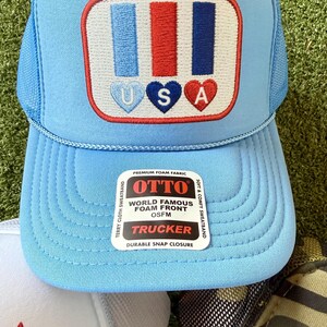 May include: Light blue trucker hat with a patriotic design. The hat features a patch with blue, red, and white stripes and the letters "USA" in heart shapes. The hat has a mesh back and a curved brim. The tag reads "OTTO WORLD FAMOUS FOAM FRONT TRUCKER".
