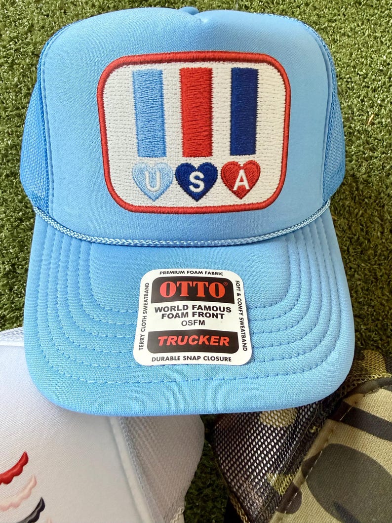 May include: Light blue trucker hat with a patriotic design. The hat features a patch with blue, red, and white stripes and the letters "USA" in heart shapes. The hat has a mesh back and a curved brim. The tag reads "OTTO WORLD FAMOUS FOAM FRONT TRUCKER".