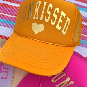 May include: A yellow trucker hat with a gold heart and the text "SUNKISSED" in gold letters.