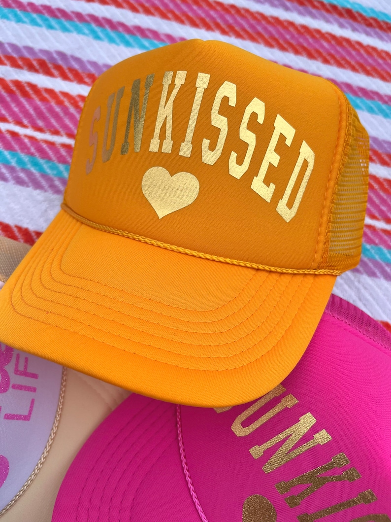 May include: A yellow trucker hat with a gold heart and the text "SUNKISSED" in gold letters.