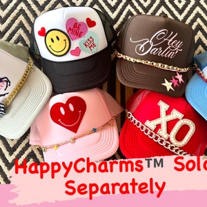May include: A collection of six trucker hats with various designs and embellishments. The hats are all different colors, including brown, pink, red, white, and blue. The designs include a smiley face, a heart, the words "Hey Darlin", "Wild Hearts Can't Be Broken", "XO", and "Love Club". The hats are all decorated with chains and other embellishments.