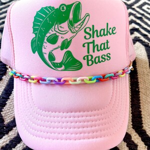 以下が含まれることがあります： ピンクのトラッカーハットに、緑のバスフィッシュのグラフィックと「Shake That Bass」の文字がプリントされています。ハットにはカラフルなチェーンのトリムが付いています。