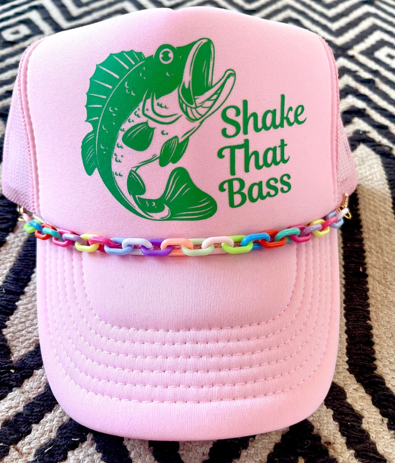 以下が含まれることがあります： ピンクのトラッカーハットに、緑のバスフィッシュのグラフィックと「Shake That Bass」の文字がプリントされています。ハットにはカラフルなチェーンのトリムが付いています。