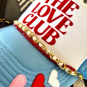 May include: A white trucker hat with a red "THE LOVE CLUB" embroidered on the front. The hat has a blue mesh back and a gold chain with red and white beads around the brim. There are three embroidered hearts on the blue front of the hat: pink, red, and white.
