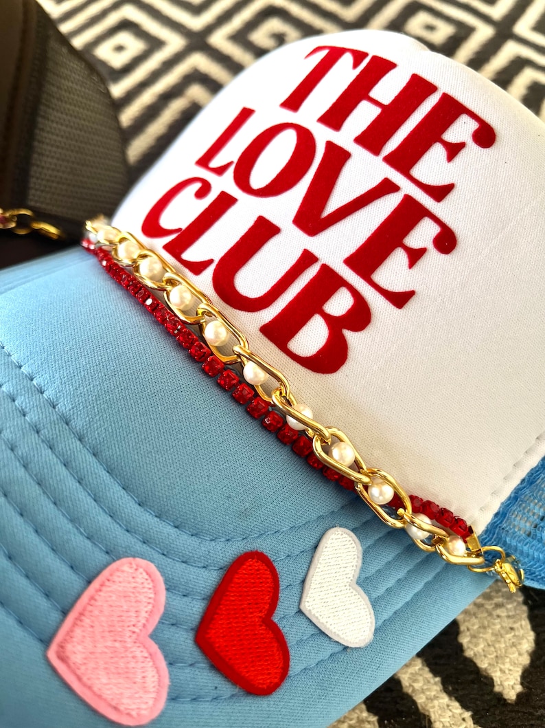 May include: A white trucker hat with a red "THE LOVE CLUB" embroidered on the front. The hat has a blue mesh back and a gold chain with red and white beads around the brim. There are three embroidered hearts on the blue front of the hat: pink, red, and white.