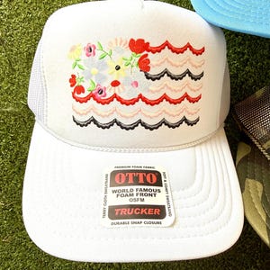 May include: White trucker hat with a floral design and wavy red, pink, and black accents. The hat features a mesh back and a curved brim. The front of the hat has the text "OTTO WORLD FAMOUS FOAM FRONT OSFM TRUCKER".