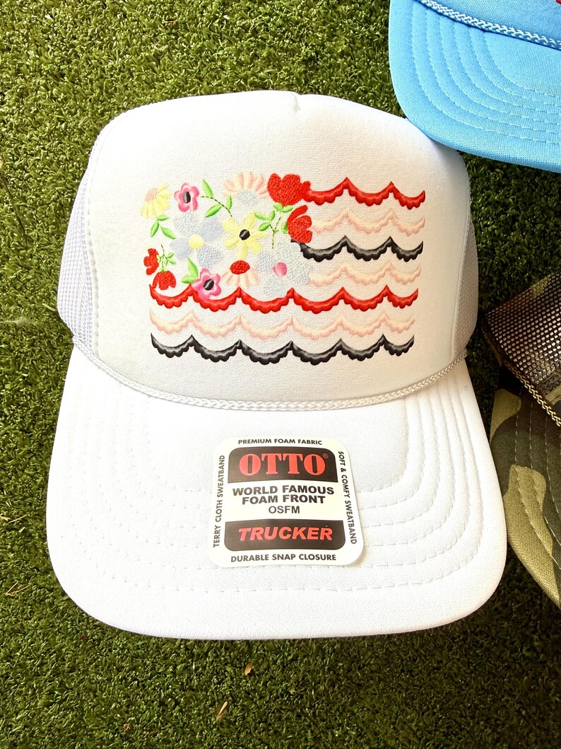 May include: White trucker hat with a floral design and wavy red, pink, and black accents. The hat features a mesh back and a curved brim. The front of the hat has the text "OTTO WORLD FAMOUS FOAM FRONT OSFM TRUCKER".