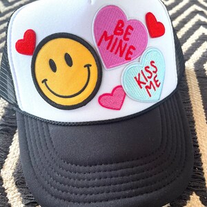 May include: A white trucker hat with a black mesh back. The hat has several patches, including a yellow smiley face, a pink heart with the text "Be Mine", a blue heart with the text "Kiss Me", and a red heart.