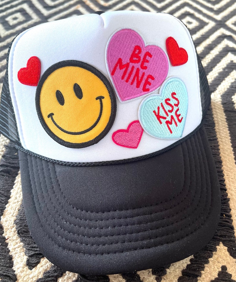 May include: A white trucker hat with a black mesh back. The hat has several patches, including a yellow smiley face, a pink heart with the text "Be Mine", a blue heart with the text "Kiss Me", and a red heart.