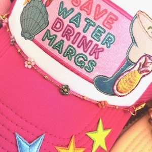May include: Pink trucker hat with a white patch that says "Save Water Drink Margs" in rainbow colors. The hat has a variety of colorful embroidered patches including a seashell, a starfish, a sun, and a margarita glass with a pineapple. The hat also has a gold chain with small flower charms.