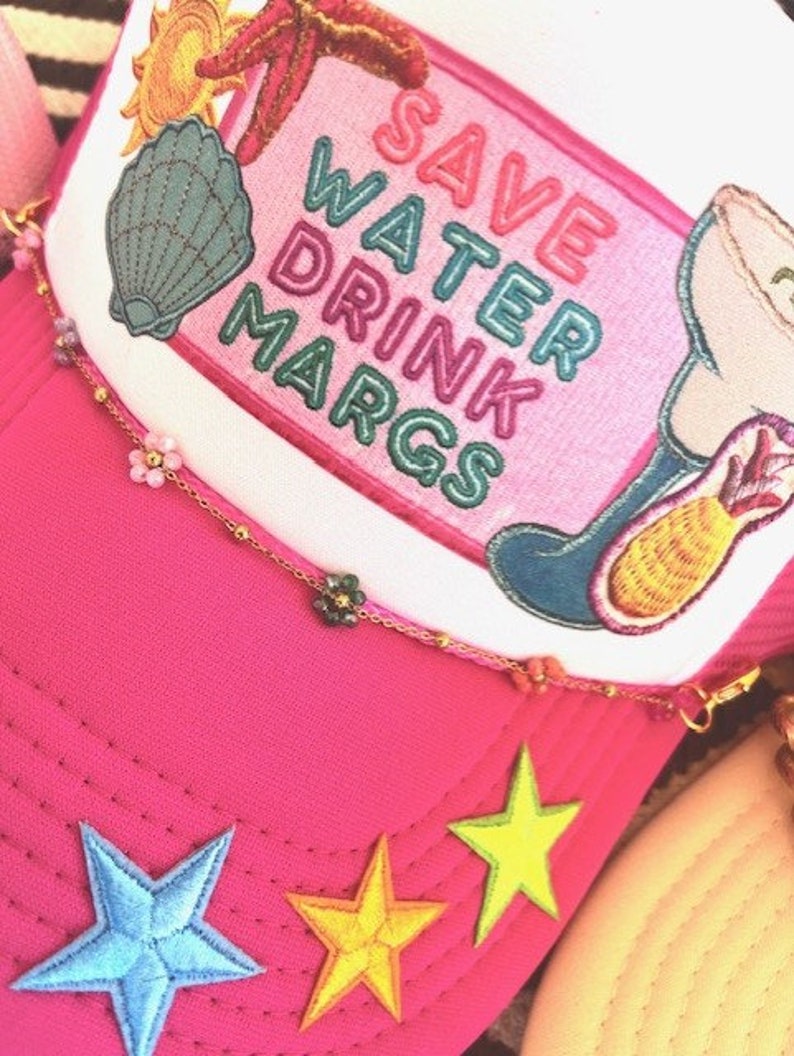 May include: Pink trucker hat with a white patch that says "Save Water Drink Margs" in rainbow colors. The hat has a variety of colorful embroidered patches including a seashell, a starfish, a sun, and a margarita glass with a pineapple. The hat also has a gold chain with small flower charms.