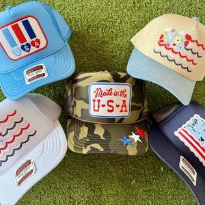 May include: A collection of trucker hats in various colors, including light blue, white, and navy. Each hat features unique embroidered designs, such as the American flag, floral patterns, and patriotic patches with "USA" text. The hats are displayed on a green surface.