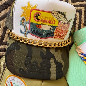 May include: A collection of trucker hats with various designs. One hat features a white front panel with a fishing-themed patch, a gold chain, and a camouflage brim. The patch includes a sun, anchor, and the words "Cotton Cordell's". Other hats have lake and fishing themes.