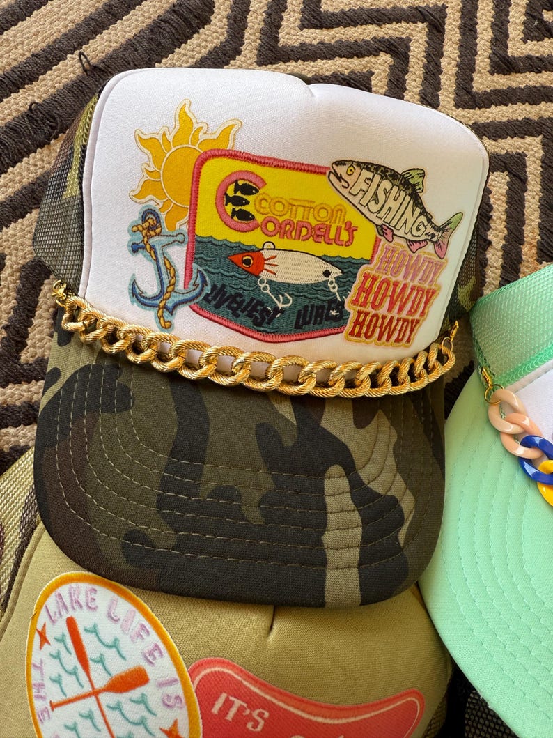 May include: A collection of trucker hats with various designs. One hat features a white front panel with a fishing-themed patch, a gold chain, and a camouflage brim. The patch includes a sun, anchor, and the words "Cotton Cordell's". Other hats have lake and fishing themes.