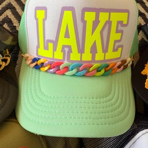 May include: A light green trucker hat with the word "LAKE" in bright yellow block letters on a white front panel. The hat features a colorful chain detail and a green mesh back.