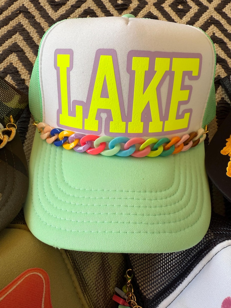 May include: A light green trucker hat with the word "LAKE" in bright yellow block letters on a white front panel. The hat features a colorful chain detail and a green mesh back.