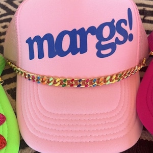 May include: Pink trucker hat with a blue "margs!" logo and a gold chain with colorful accents.