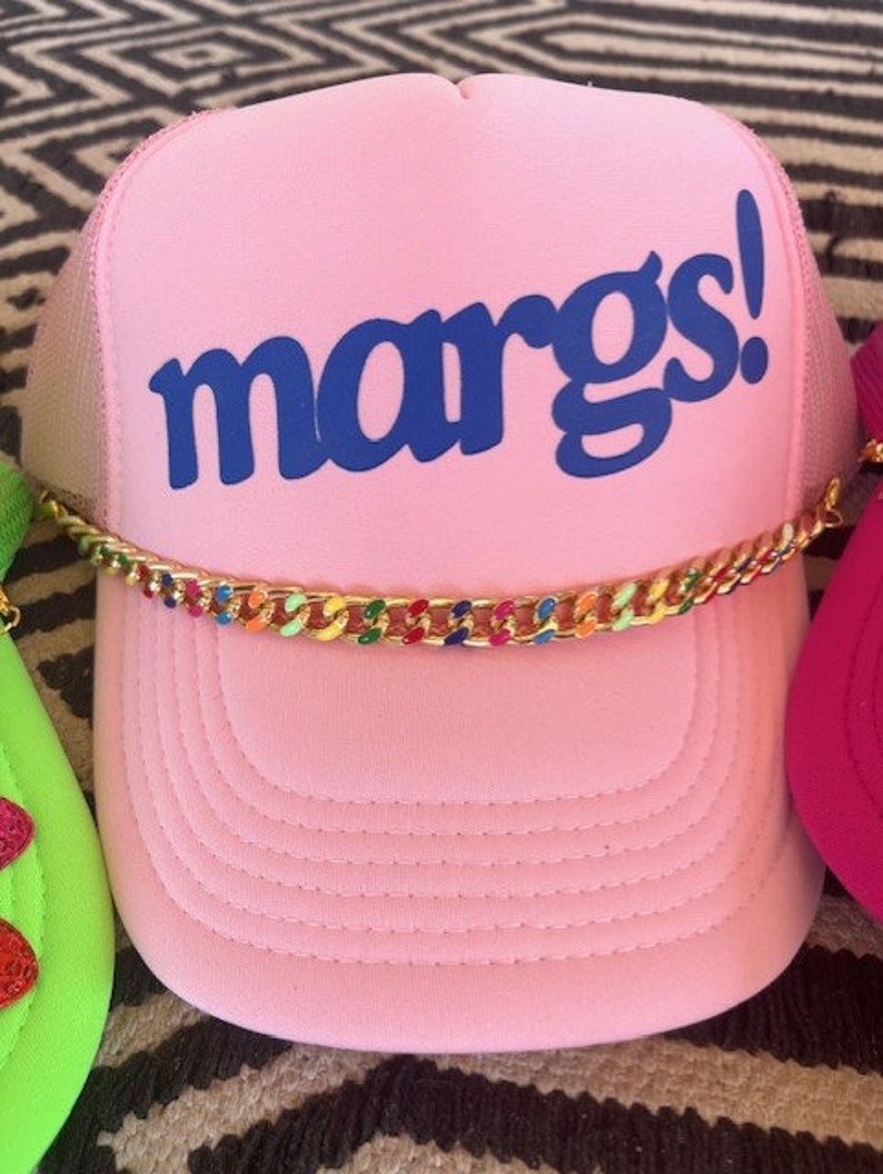 May include: Pink trucker hat with a blue "margs!" logo and a gold chain with colorful accents.