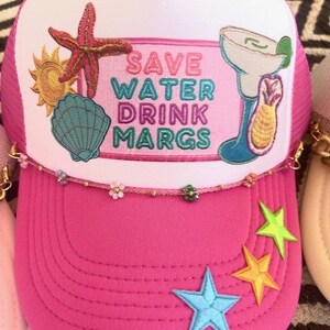 May include: Pink trucker hat with a white mesh back and a white front panel featuring a colorful embroidered design that says "Save Water Drink Margs". The design includes a starfish, a seashell, a sun, and a margarita glass with a lime wedge. The hat has a gold chain trim and three embroidered stars on the pink brim.