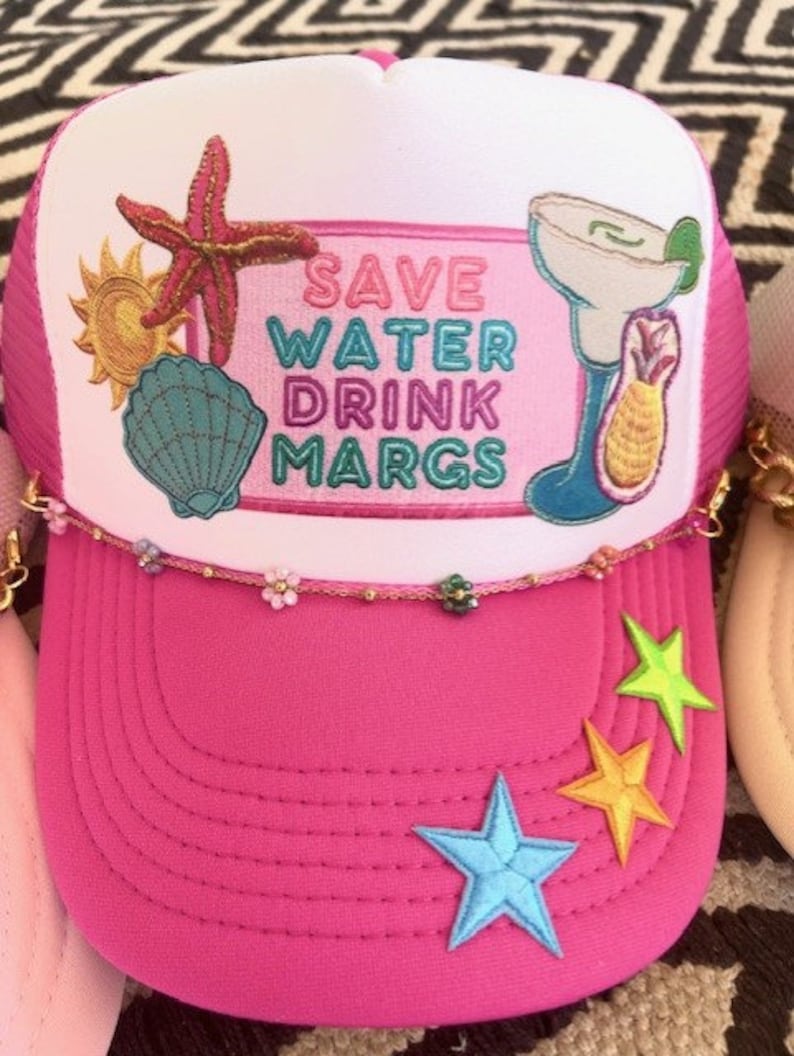 May include: Pink trucker hat with a white mesh back and a white front panel featuring a colorful embroidered design that says "Save Water Drink Margs". The design includes a starfish, a seashell, a sun, and a margarita glass with a lime wedge. The hat has a gold chain trim and three embroidered stars on the pink brim.