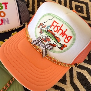 May include: Orange and white trucker hat with a fishing-themed patch. The patch features a green and red fish with a worm on a hook, and the word "Fishing". The hat has a gold chain and a gingham bow.