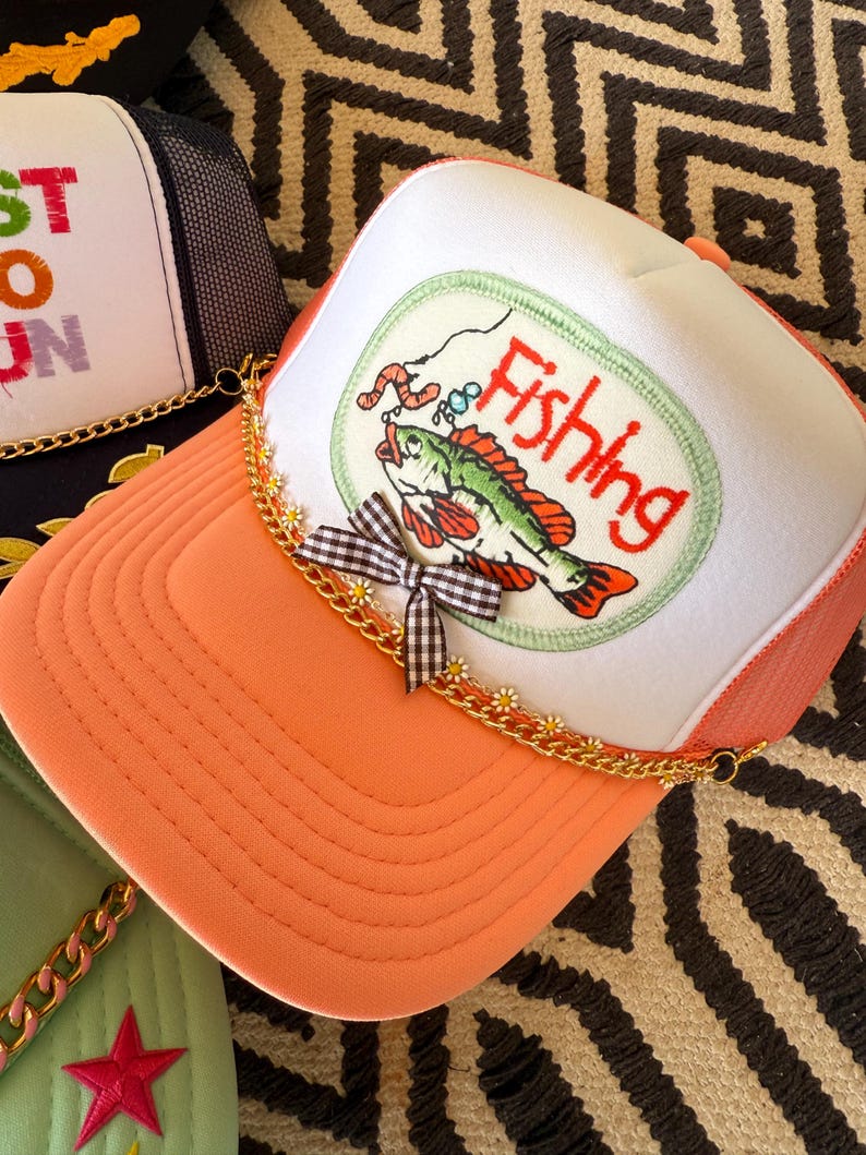 May include: Orange and white trucker hat with a fishing-themed patch. The patch features a green and red fish with a worm on a hook, and the word "Fishing". The hat has a gold chain and a gingham bow.