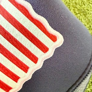 May include: Close-up of a navy blue hat with a patriotic design. The design features red and white stripes, with a white border. The hat is made of a textured fabric and is set against a green background.