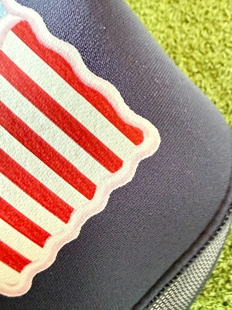 May include: Close-up of a navy blue hat with a patriotic design. The design features red and white stripes, with a white border. The hat is made of a textured fabric and is set against a green background.