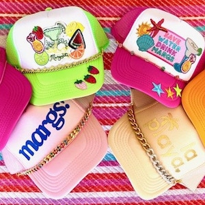 May include: Six colorful trucker hats with various designs and phrases, including "Sunkissed", "Marg's", "Save Water Drink Margs", and "Oaxaca".