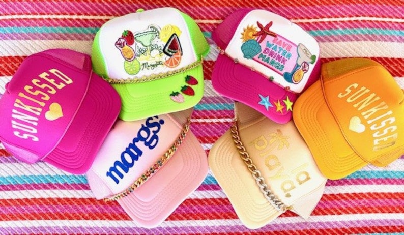 May include: Six colorful trucker hats with various designs and phrases, including "Sunkissed", "Marg's", "Save Water Drink Margs", and "Oaxaca".