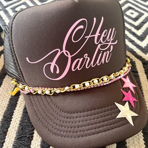 May include: A black trucker hat with a pink "Hey Darlin'" design. The hat has a gold chain with white pearls and pink and white stars attached.