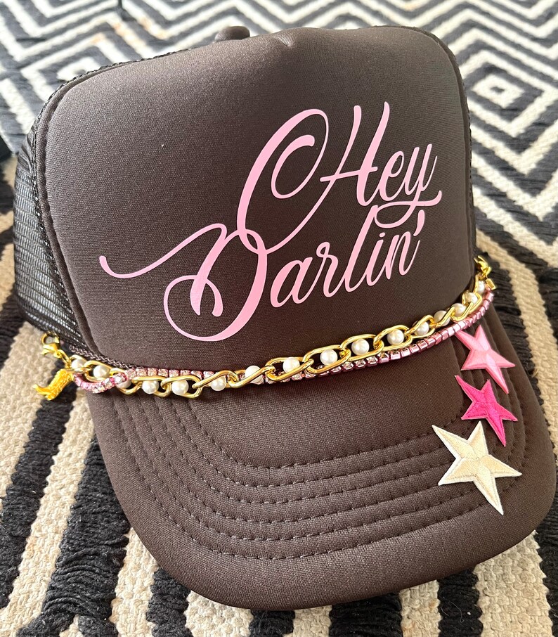 May include: A black trucker hat with a pink "Hey Darlin'" design. The hat has a gold chain with white pearls and pink and white stars attached.