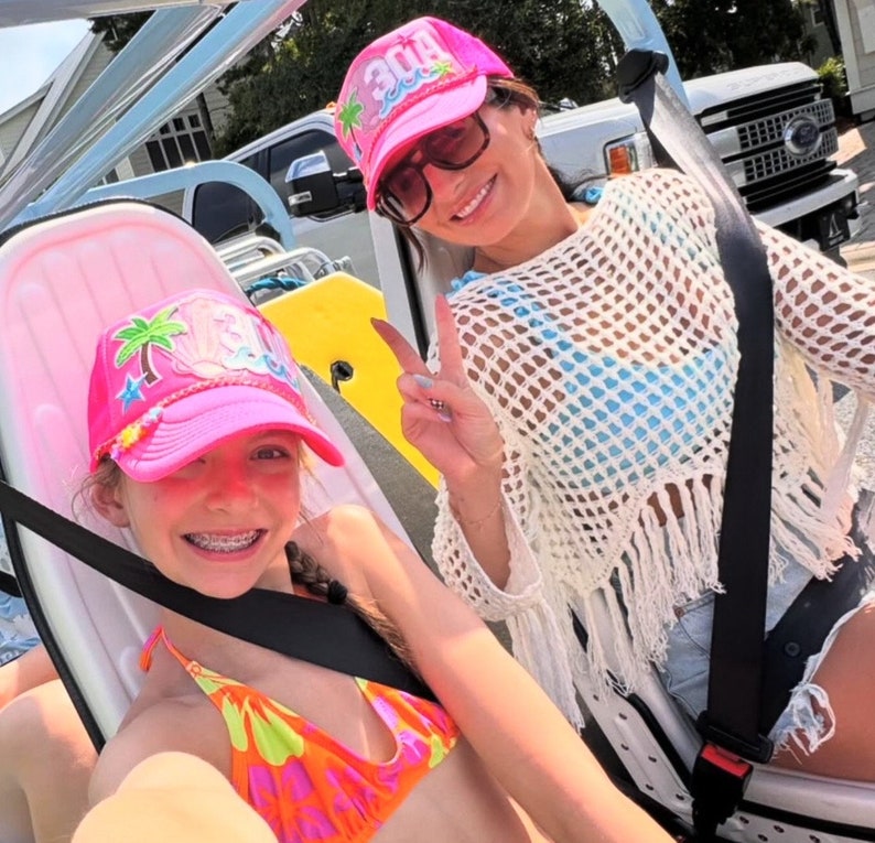 Op de afbeelding: Twee mensen dragen roze en witte hoeden met het woord "30A" erop. De persoon vooraan draagt een roze en oranje bikini top en een zwarte harnas. De persoon achteraan draagt een wit gehaakt top met een blauwe en witte bikini top eronder.