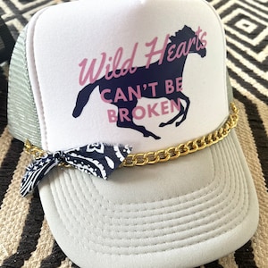 May include: A white trucker hat with a navy blue horse and the text "Wild Hearts Can't Be Broken" in pink lettering. The hat has a gold chain and a blue bandana tied around it.