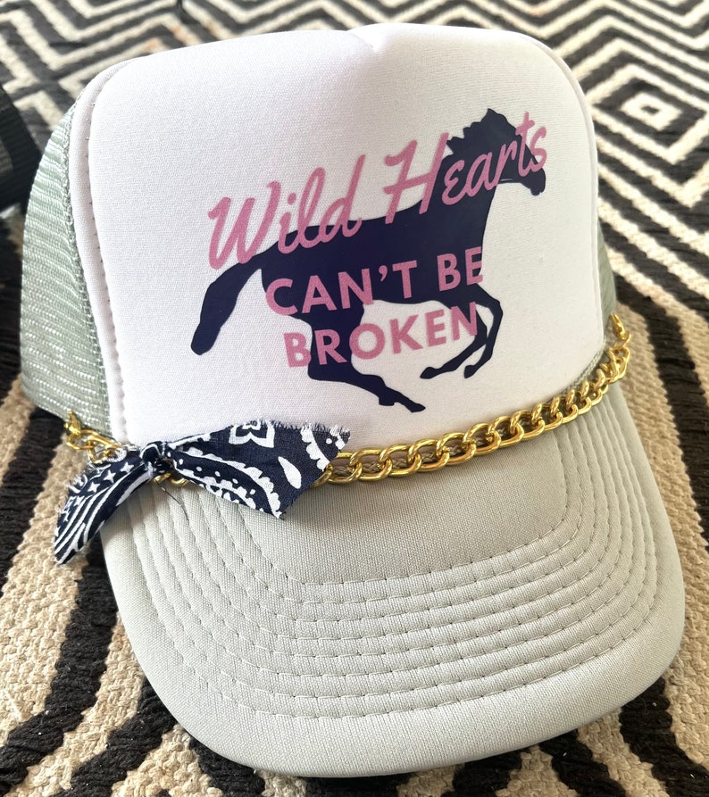 May include: A white trucker hat with a navy blue horse and the text "Wild Hearts Can't Be Broken" in pink lettering. The hat has a gold chain and a blue bandana tied around it.