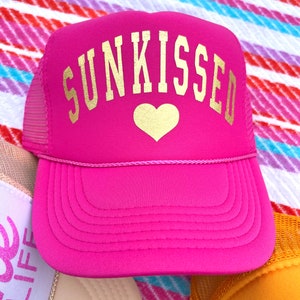 May include: Pink trucker hat with a gold mesh back and a gold embroidered heart and the word "SUNKISSED" on the front.