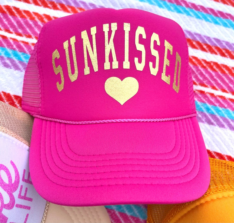 May include: Pink trucker hat with a gold mesh back and a gold embroidered heart and the word "SUNKISSED" on the front.