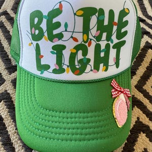 May include: A green and white trucker hat with the words "Be the Light" in green letters with colorful Christmas lights strung around the letters. A pink and white Christmas ornament with a red bow is attached to the hat.