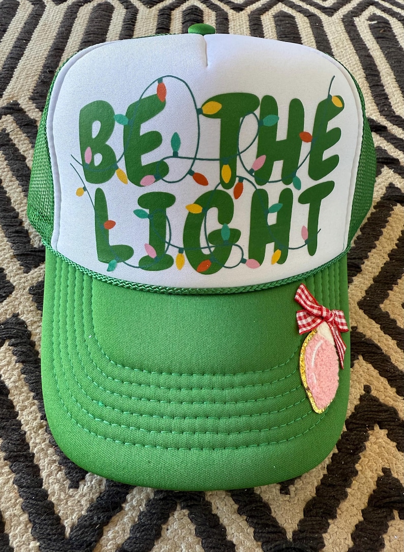 May include: A green and white trucker hat with the words "Be the Light" in green letters with colorful Christmas lights strung around the letters. A pink and white Christmas ornament with a red bow is attached to the hat.