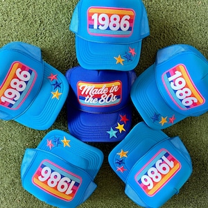 May include: A collection of blue trucker hats with the year "1986" displayed on the front in a retro design. Some hats feature star accents. One hat reads "Made in the 80s". The hats are arranged on a green surface.